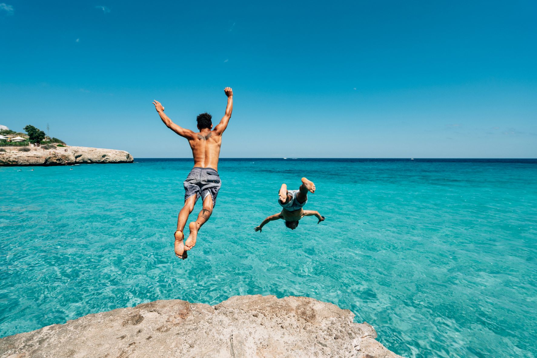 Two men are leaping into crystal-clear ocean waters. There's not a cloud in the sky.